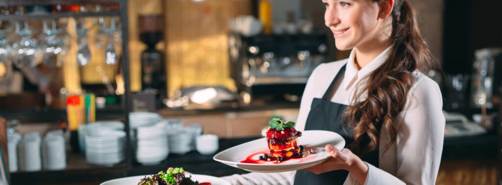 Une jeune serveuse en formation portant un tablier noir et deux assiettes de desserts